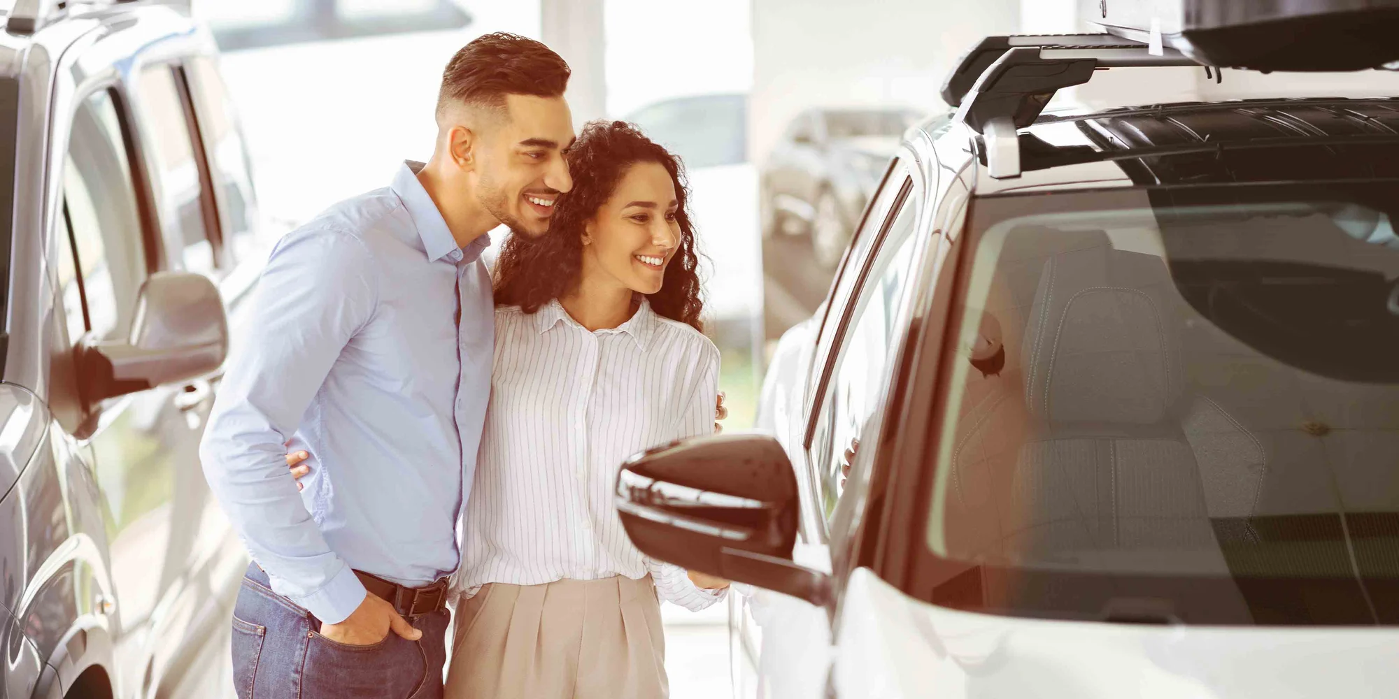 A young man and woman stand with their arms around each other as they look into the inside of a vehicle in a vehicle showroom.