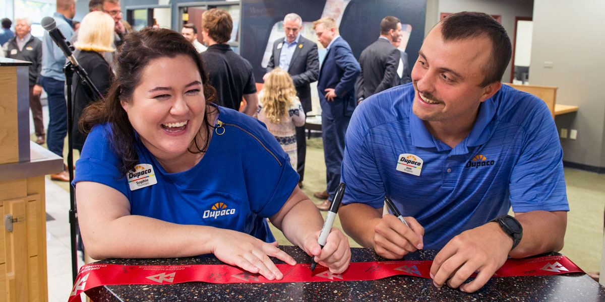 Dupaco’s Sophie Thomsen and Jon Nugent sign their names on the red ribbon from the Sept. 27 ribbon-cutting ceremony at the Mineral Point Road branch in Madison, Wis. (K. Wolf photo)