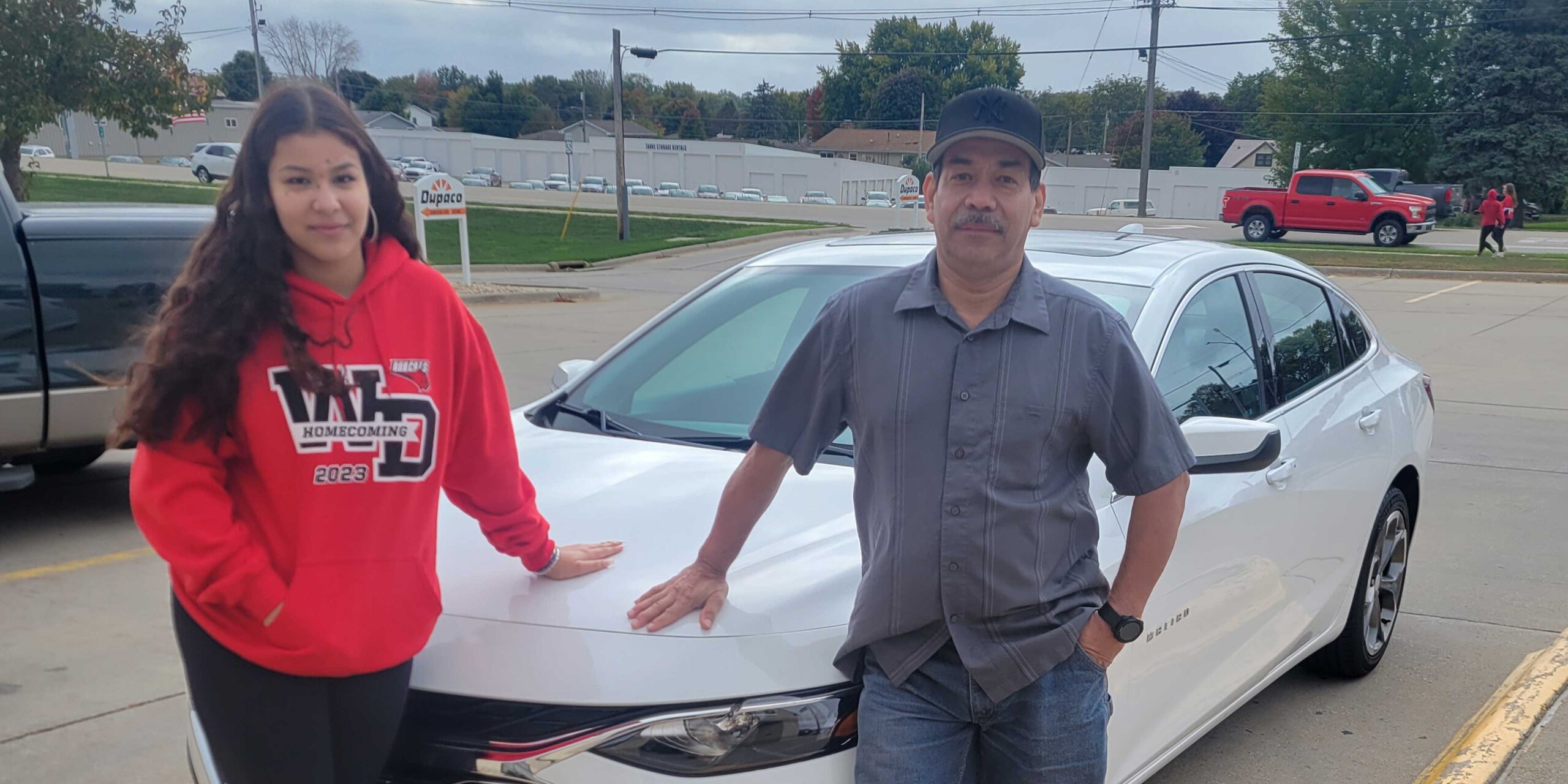 A father and his daughter standing in front of their white car outside in a parking lot.