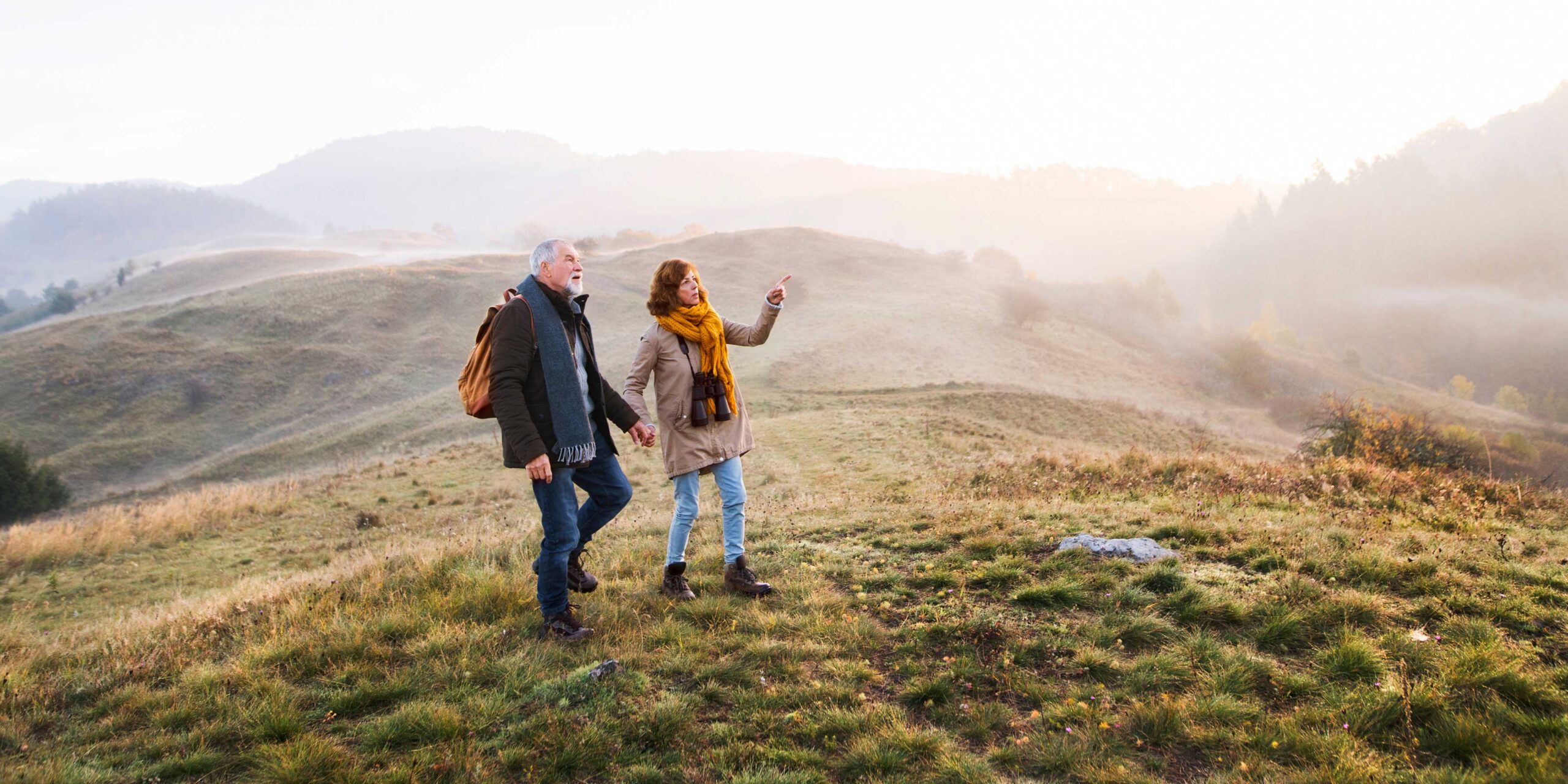 Middle-aged couple happily exploring a vast grassland, hand in hand, surrounded by the beauty of nature, with hills and foggy white skies in the background.